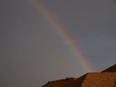 [The bow's arc extends from behing a shingled rooftop in the lower right to a section of sky in the upper left. The sky is somewhat dark which mutes the colors of the rainbow, but violet, green, yellow, and orange-red are visible from left to right.]
